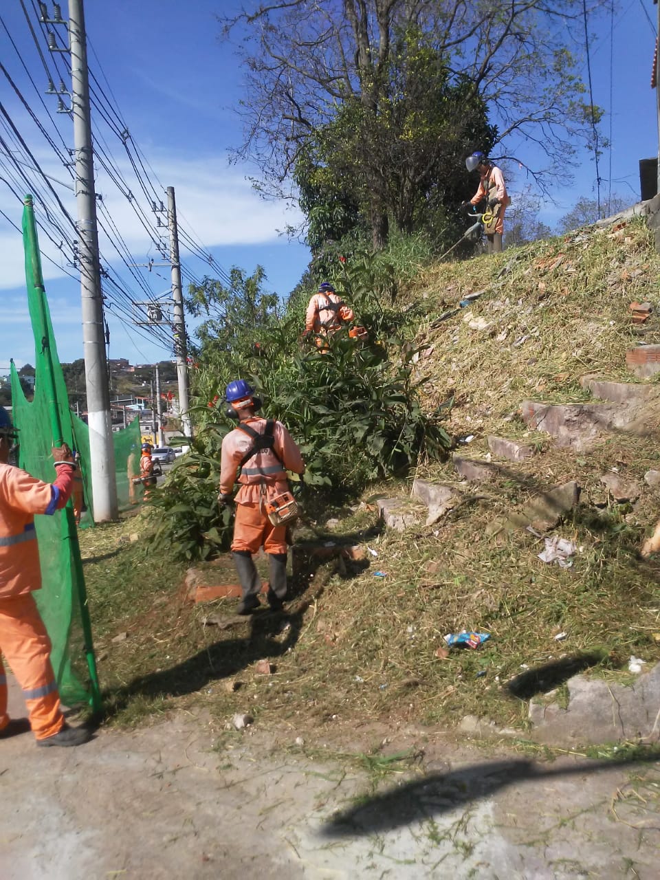 Quatros funcionários, vestidos com o uniforme da Prefeitura e munidos de equipamentos, como tela de proteção e roçadeira, realizando poda de grama num relevo inclinado.